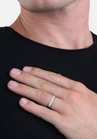 Srebrny pierścionek o gładkim, zaokrąglonym kształcie, spoczywający na dłoni. Charakteryzuje się subtelnym połyskiem, minimalistycznym designem, umieszczony na czarnej koszuli.