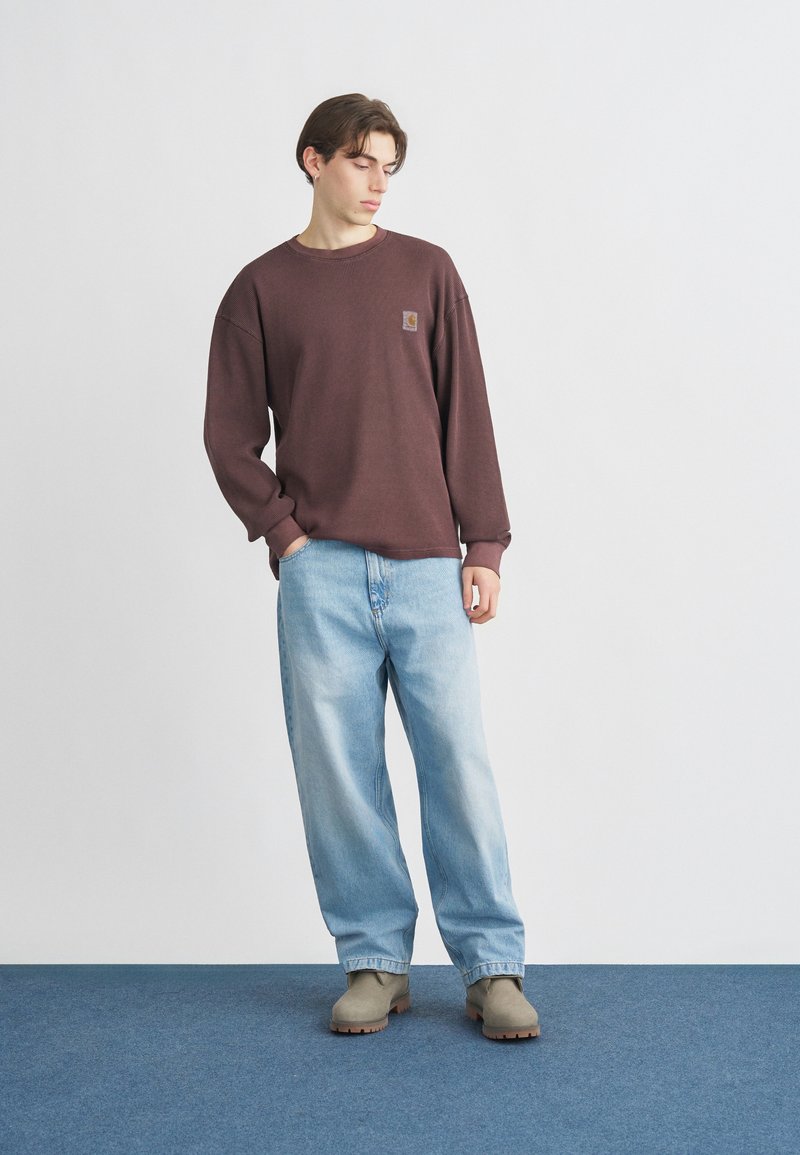 Brun ribstrikket sweatshirt med lille logo, kombineret med lyseblå løse jeans og beige støvler. Ensfarvet baggrund, stående på blåt tæppe.