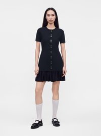 Femme debout dans une robe noire à manches courtes avec des boutons sur le devant et un ourlet plissé, portant des chaussettes blanches montantes et des chaussures noires.