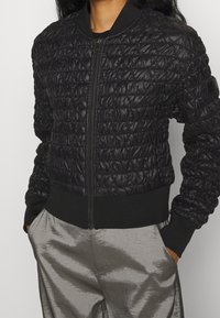 Osoba v černé prošívané bomber bundě s vytáhnutým zipem a kapsami, s rukama v kapsách, na bílém pozadí.