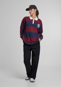 Červeno-modro pruhované ragbyové tričko s bielym golierom, s logom na náplete. V kombinácii s čiernymi nohavicami voľného strihu a bielymi teniskami.