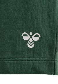Grön bomullstyg med en subtil textur har en vit bi-logotyp vid den nedre kanten, omgiven av en söm.