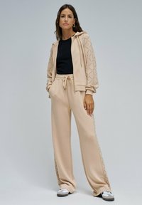 Sudadera beige con capucha y mangas estampadas, combinada con pantalones anchos a juego. Camiseta negra debajo. Calzado casual, diseño simple.