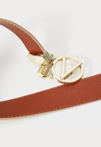 Brūns auduma jostas ar zelta metāla sprādzi un apaļu logo, kurā ir V dizains. Tekstūra ir gluda ar delikātu spīdumu.