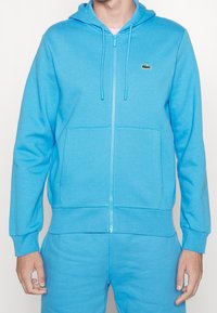 Světle modrá mikina na zip vyrobená z měkké tkaniny. Obsahuje přední kapsu a kapuci na šňůrku. Na hrudi je malé vyšívané logo.