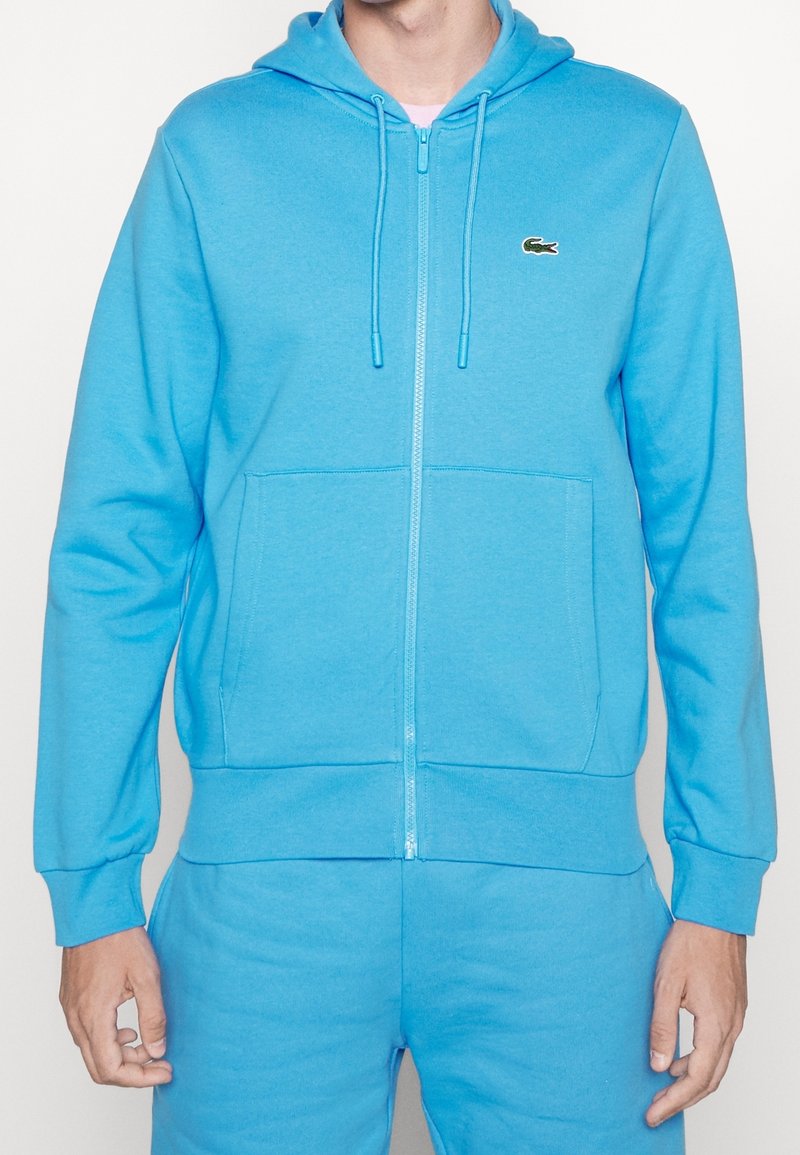 Světle modrá mikina na zip vyrobená z měkké tkaniny. Obsahuje přední kapsu a kapuci na šňůrku. Na hrudi je malé vyšívané logo.