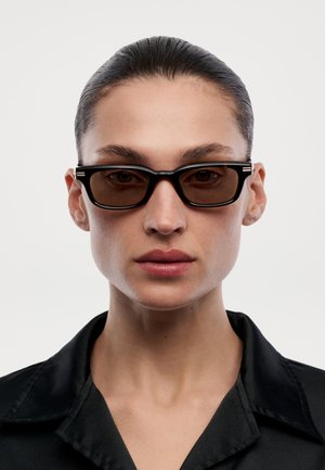 Giovane donna con capelli tirati indietro, che indossa occhiali da sole rettangolari scuri e una camicia nera con colletto, che guarda dritto davanti a sé.