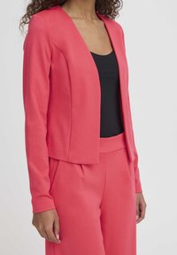 Veste rose à manches longues en tissu lisse avec un col en V, portée sur un haut noir, assortie à un pantalon rose assorti.