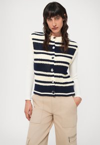 Colete de malha em riscas azul-marinho e creme, com decote redondo e fecho à frente com botões. Combinado com calças cargo bejes, realçando elementos de múltiplas texturas.