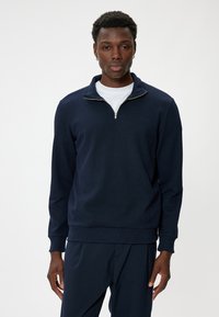 Sudadera de media cremallera azul marino con textura canalé y cremallera plateada, combinada con una camiseta blanca de cuello redondo y pantalones azul marino.