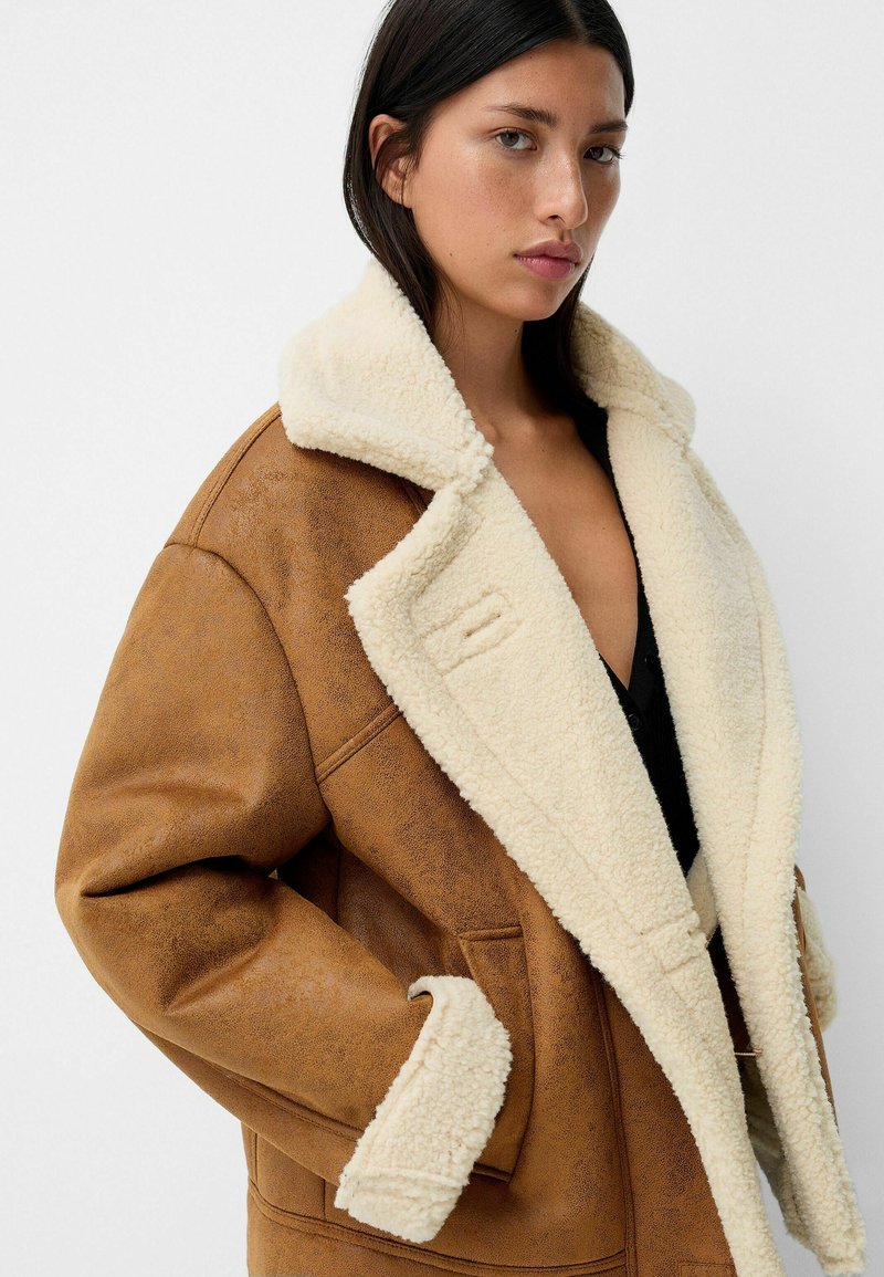 Abrigo de shearling marrón con un gran cuello crema, que cuenta con bolsillos frontales y un exterior texturizado. El abrigo tiene un corte relajado.
