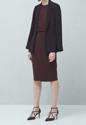 Czarna marynarka na burgundowej sukience z węzłem w talii. Czarne szpilki z paskami wokół kostki i ćwiekami.