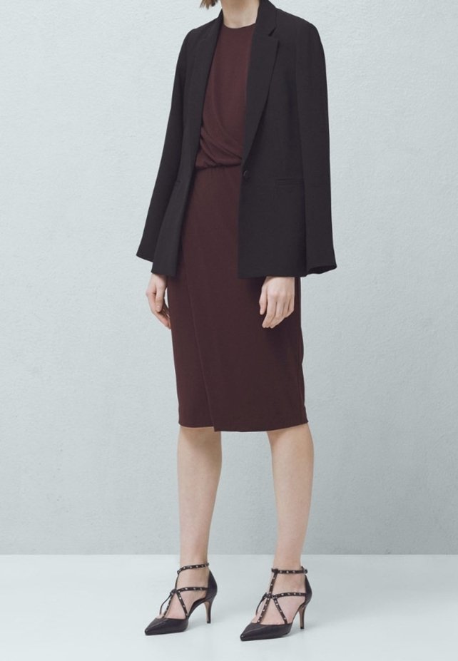 Czarna marynarka na burgundowej sukience z węzłem w talii. Czarne szpilki z paskami wokół kostki i ćwiekami.