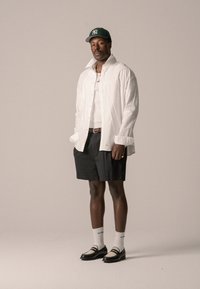 Camicia bianca a pulsanti, pantaloni corti neri, cappellino da baseball verde, collana d'oro, scarpe nere e crema, calze bianche con logo, in piedi su uno sfondo neutro.