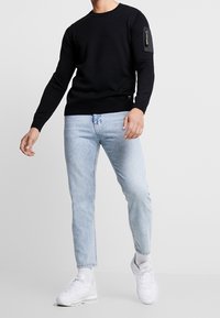 Sweatshirt en maille noire avec un col rond et des manches texturées, équipé d'une poche zippée sur le bras gauche. Assorti à un jean bleu clair et des baskets blanches.