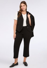 Set di pantaloni e gilet neri su misura abbinati a una t-shirt bianca; i pantaloni hanno un taglio cropped, con fronte piatto e passanti per la cintura. Mocassini neri completano il look.