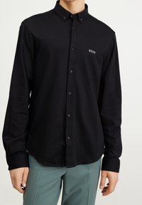Schwarzes Hemd mit Knopfleiste und klassischem Kragen, mit einem dezenten Logo auf der Brust, aus glattem Stoff gefertigt und mit langen Ärmeln.
