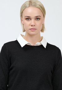 Donna bionda con capelli corti che indossa una camicia bianca con colletto sotto un maglione nero a girocollo, rivolta in avanti con un'espressione neutra.
