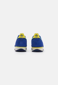 Blauwe suede sneakers met gele accenten, met een textuur op de hak en een witte tussenzool. De achterkant toont het Adidas-logo.