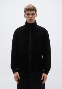 Μαύρο σακάκι από σουέντ με ψηλό γιακά, ολόκληρο φερμουάρ μπροστά και πλαϊνές τσέπες· διαθέτει μαλακή υφή και άνετη εφαρμογή.