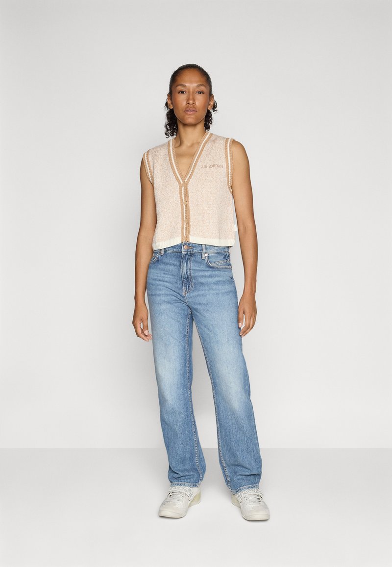 Beige strickweste ohne Ärmel mit V-Ausschnitt und vorderem Knopfverschluss, kombiniert mit hellblauen geraden Jeans und weißen Sneakers.