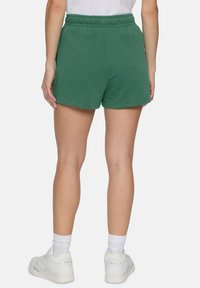 Pantaloni corti in cotone verde con vita elasticizzata, tasche laterali e vestibilità comoda. Indossati con scarpe sportive bianche e calzini alti.