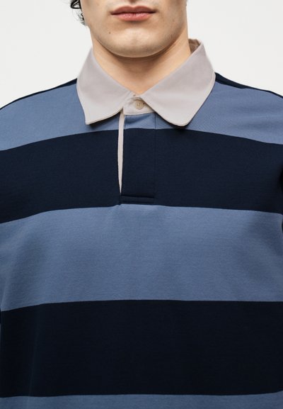 Homme portant un maillot de rugby à larges rayures horizontales bleu et noir avec un col boutonné beige clair, vu du cou à la poitrine.