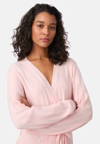 Jasnoróżowy sweter kardigan z dzianiny, z dekoltem w szpic, fakturą prążkowaną i otwartym splotem. Posiada długie rękawy oraz wiązanie w pasie.