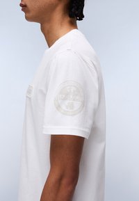 Maglietta bianca in cotone con maniche corte, caratterizzata da un logo leggermente in rilievo sulla manica sinistra e un'etichetta in tessuto sul lato frontale.