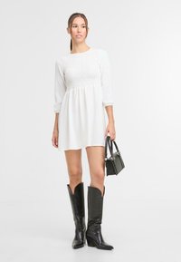 Witte jurk met tekstuur en smock-bustier en lange mouwen. Zwarte leren kniehoge laarzen met spitse neuzen. Kleine zwarte handtas.
