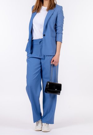 Vrouw draagt een blauw damespak en witte sneakers, met een kleine zwarte handtas met kettingriem, tegen een witte achtergrond.