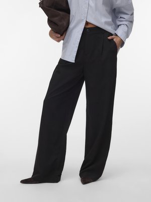 Άτομο που φοράει άνετο μαύρο παντελόνι και ανοιχτό γαλάζιο πουκάμισο, κρατώντας μια σκούρα καφέ τσάντα, με το ένα χέρι στην τσέπη.