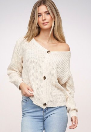 Junge Frau mit langen blonden Haaren, die eine cremefarbene Strickjacke mit Schultern aus dem Kragen und Knöpfen sowie hellblaue Jeans trägt, steht vor einem schlichten Hintergrund.