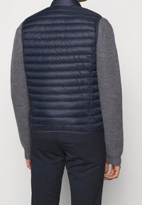 Navy puffer vest with horizontal stitching, high collar, and lightweight material. Paired with a textured gray long-sleeve sweater.