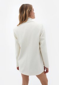 Blazer bianco su misura con design strutturato, maniche lunghe, retro leggermente allungato e texture liscia. Foderato con una leggera lucentezza.