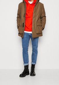Brown parka with a hood, zippered front, and large pockets layered over a bright red hoodie, paired with blue jeans and black lace-up boots.