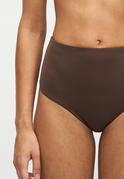 Gros plan sur la région médiane d’une personne portant un sous-vêtement marron taille haute, montrant une partie du bras et du torse sur un fond uni.