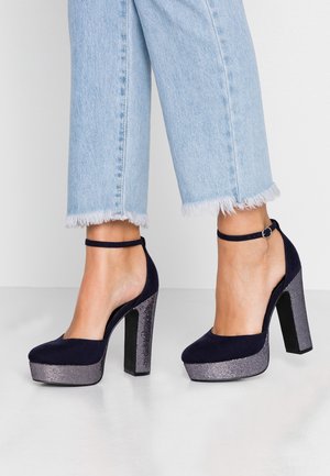 Marineblauwe suède hoge hakken met een plateauzool en een metallic textuur op de hak. Voorzien van een enkelbandje en een open achterkant. Spijkerbroek met rafelige zoom.
