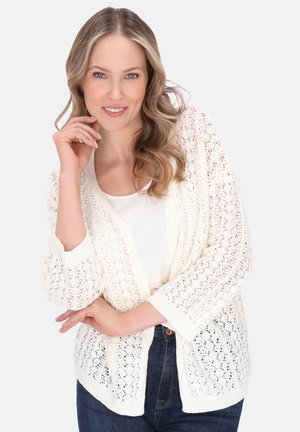 Häkelstrick-Kardigan in Creme mit gerippten Bündchen, weichem Textil und lockerer Passform. Über einem weißen Oberteil getragen und mit dunklen Denim-Jeans kombiniert.
