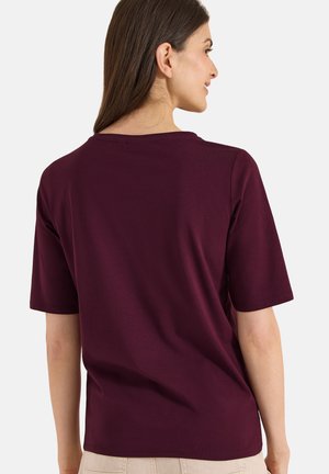 Kobieta z długimi brązowymi włosami w jednolitej bordowej koszulce z krótkim rękawem, lekko obrócona bokiem, pokazująca plecy.