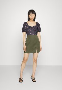 Top preto florido com mangas bufantes e decote arredondado, combinado com uma saia mini verde com fenda lateral e textura suave.