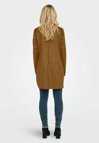Manteau long en suède synthétique marron avec une coupe droite, associé à un jean skinny bleu et des bottines noires, vu de dos. Cheveux longs et ondulés.