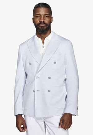 Mężczyzna z krótkimi włosami i brodą, ubrany w jasnoniebieską dwurzędową marynarkę na białą koszulę na zamek i białe spodnie, stojący na białym tle.