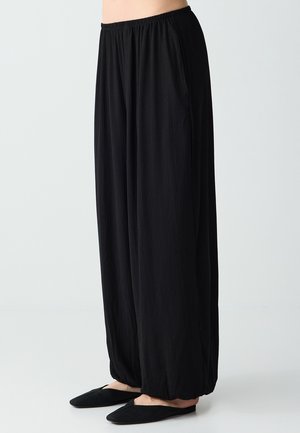 Luźne czarne plisowane spodnie z elastycznym pasem, noszone z czarnymi wsuwanymi płaskimi butami, pokazane na osobie od talii do stóp.