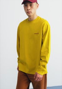Long-sleeved yellow cotton t-shirt with a crew neck and embroidered logo. Paired with brown pants, against a neutral background.