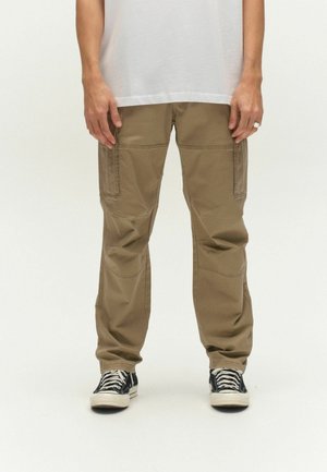 Person iført beige cargobukser og sorte høje sneakers, hænder afslappet ved siderne, ensfarvet hvid baggrund.