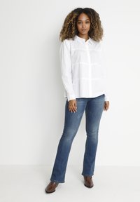 White button-up shirt with pleated details, paired with dark blue straight-leg jeans and brown ankle boots. Simple, clean design.