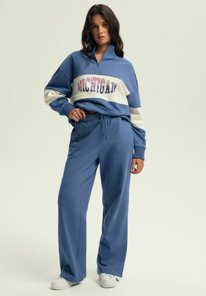 Jasnoniebieski sweatshirt z zamkiem błyskawicznym w ćwiartkę oraz napisem "MICHIGAN"; w zestawie z luźnymi spodniami i białymi sneakersami. Miękka tekstura tkaniny.