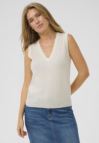 Haut sans manches crème avec un décolleté en V, fabriqué à partir d'un tissu tricot léger. Texture lisse, design ajusté. Porté avec une jupe en denim bleu.