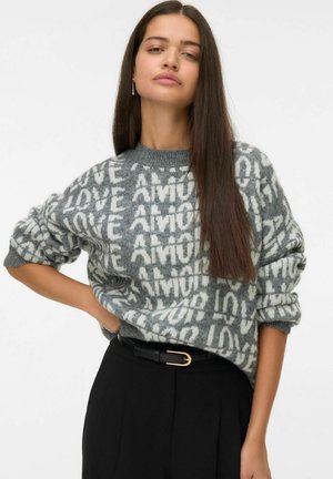 Femme avec de longs cheveux bruns portant un pull gris avec un motif de texte blanc "LOVE" et un pantalon noir avec une ceinture noire à boucle en or.